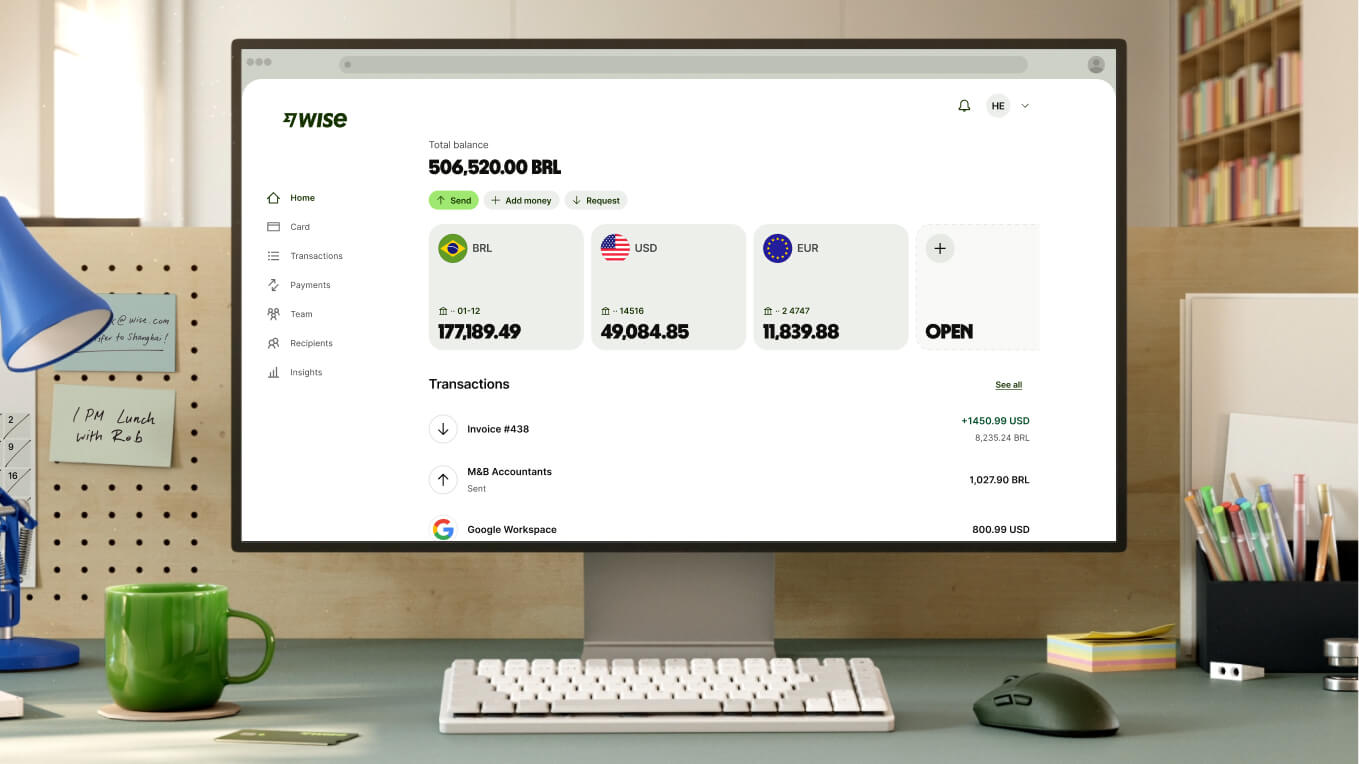 A close-up shot of a modern workspace shows a white computer monitor displaying the Wise dashboard. The screen shows a total balance of "506,520.00 BRL" and three main currency accounts: BRL with "177,189.49," USD with "49,084.85," and EUR with "11,839.88," followed by an option to open a new account. Recent transactions are listed below. In front of the monitor is a compact white keyboard, a dark grey mouse, and a bright green ceramic mug.