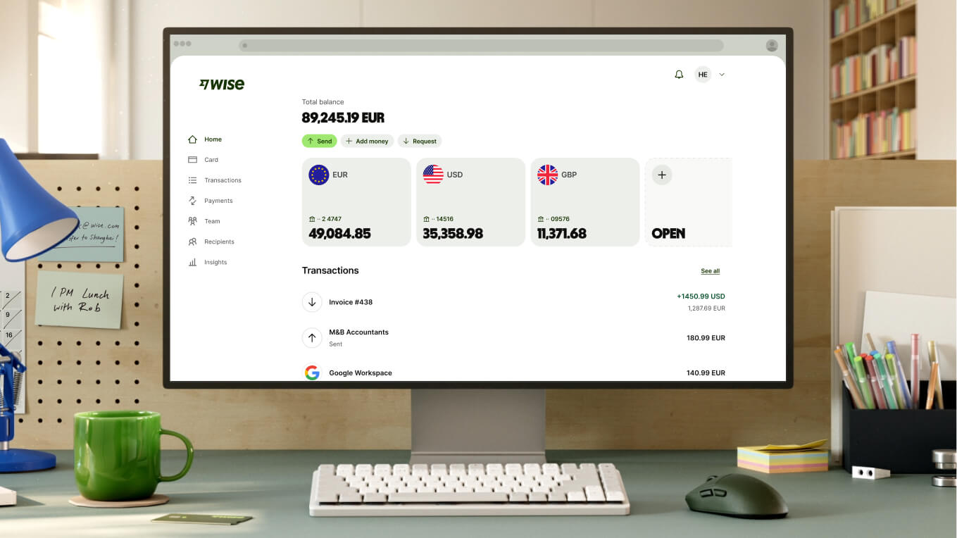 O fotografie de aproape a unui spațiu de lucru modern arată un monitor alb de computer care afișează pagina principală Wise. Ecranul afișează un sold total de 89.245,19 EUR și trei conturi valutare principale: EUR cu 49.084,85, USD cu 35.358,98 și GBP cu 11.371,68, urmat de o opțiune de deschidere a unui cont nou. Tranzacțiile recente sunt enumerate mai jos. În fața monitorului se află o tastatură albă compactă, un mouse gri închis și o cană ceramică verde strălucitor.