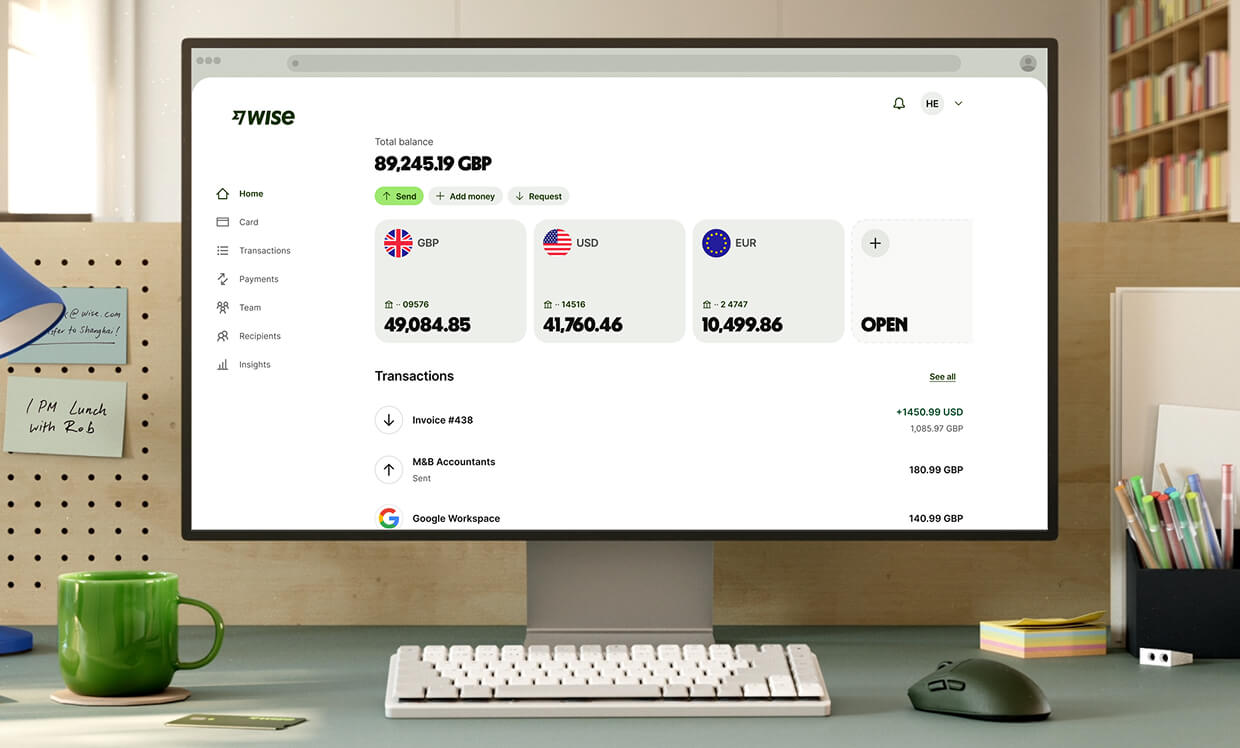 Eine Nahaufnahme eines modernen Arbeitsplatzes zeigt einen weißen Computermonitor, auf dem das Wise-Dashboard angezeigt wird. Der Bildschirm zeigt ein Gesamtguthaben von "89.245,19 GBP" und drei Hauptwährungskonten: GBP mit "49.084,85", USD mit "41.760,46" und EUR mit "10.499,86", gefolgt von einer Option zur Eröffnung eines neuen Kontos. Darunter werden die letzten Transaktionen aufgelistet. Vor dem Monitor befinden sich eine kompakte weiße Tastatur, eine dunkelgraue Maus und eine leuchtend grüne Keramiktasse.