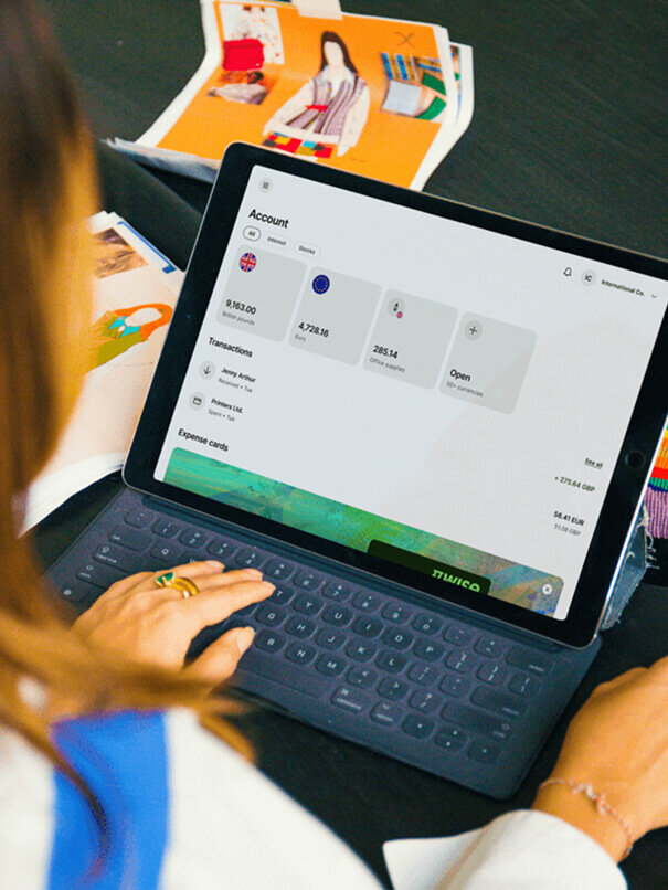 A person, seen from over their shoulder, is typing on a tablet. The tablet screen displays an "Account" dashboard with balances, transactions, and "Expense cards."